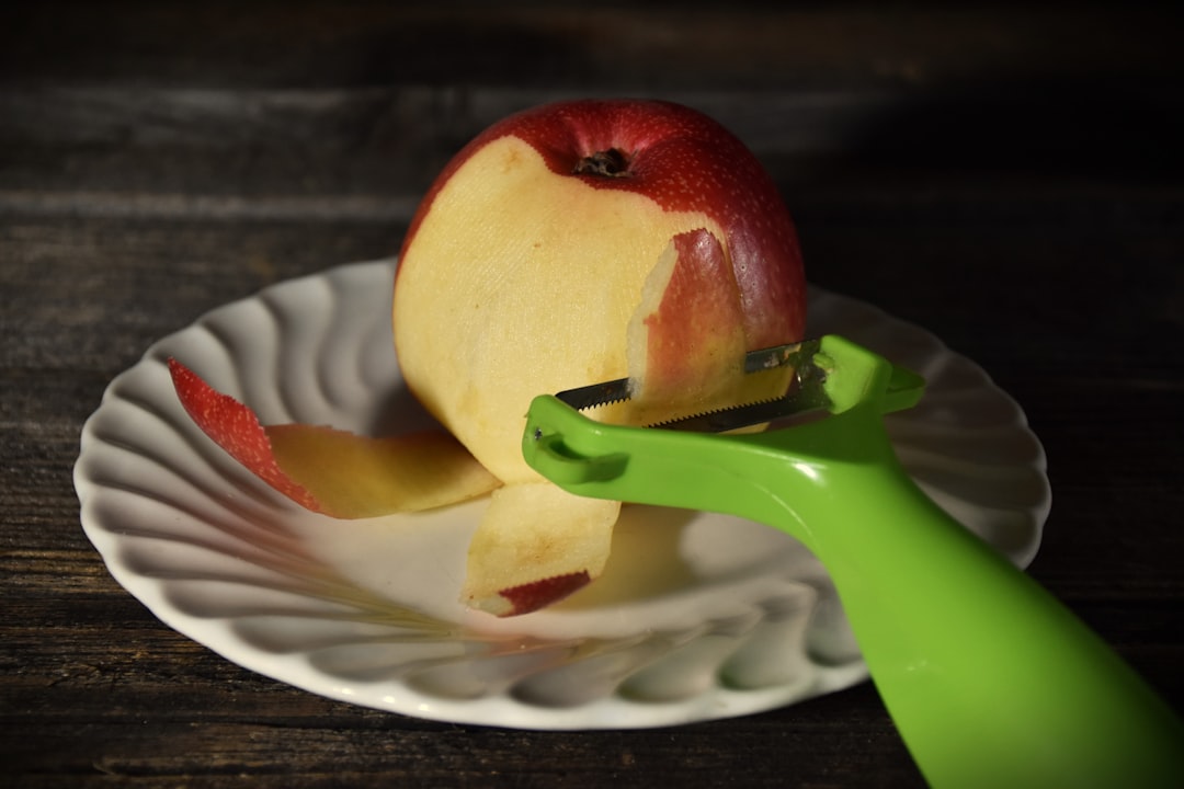 A red apple being peeled with a vegetable peeler over a white plate